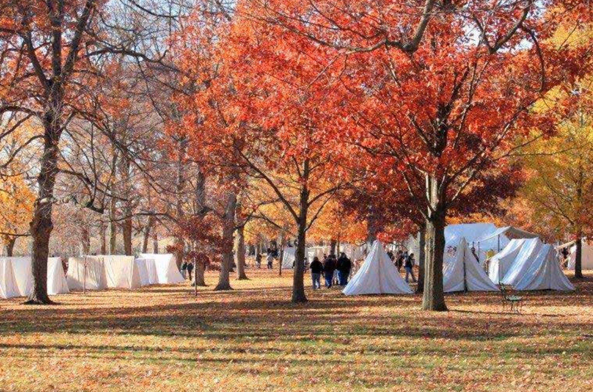 a fall foliage shot of an open field with some trees and several white tents.