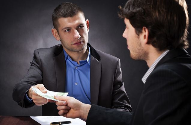 Man handing another man money over a desk they are both seated at